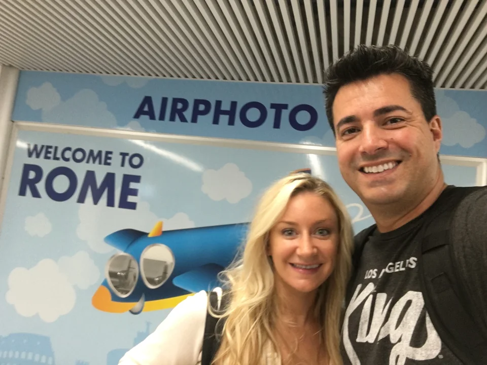 Alex and Kristin smiling together beneath an airport welcome sign in Rome